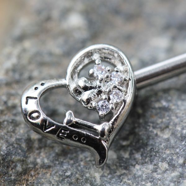 A silver-toned heart-shaped nipple barbell with a cluster of clear gemstones on one side and the word 'LOVE' engraved on the other, resting on a textured gray stone surface.

