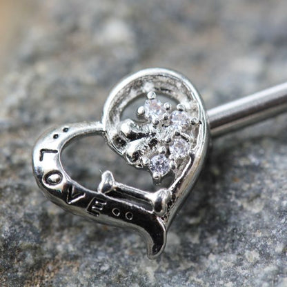A silver-toned heart-shaped nipple barbell with a cluster of clear gemstones on one side and the word 'LOVE' engraved on the other, resting on a textured gray stone surface.

